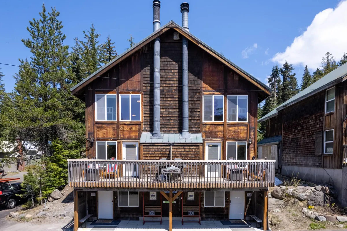 The image showcases the inviting exterior of a spacious town house nestled in the mountains, featuring a welcoming wooden deck perfect for gatherings, charming chimneys, and expansive windows that hint at cozy interiors. The deck overlooks the surrounding natural beauty, with the warm glow of sunset casting a golden hue and a fire pit ready for evening relaxation. Visible signage reinforces the home's unique identity and connection to the local community.