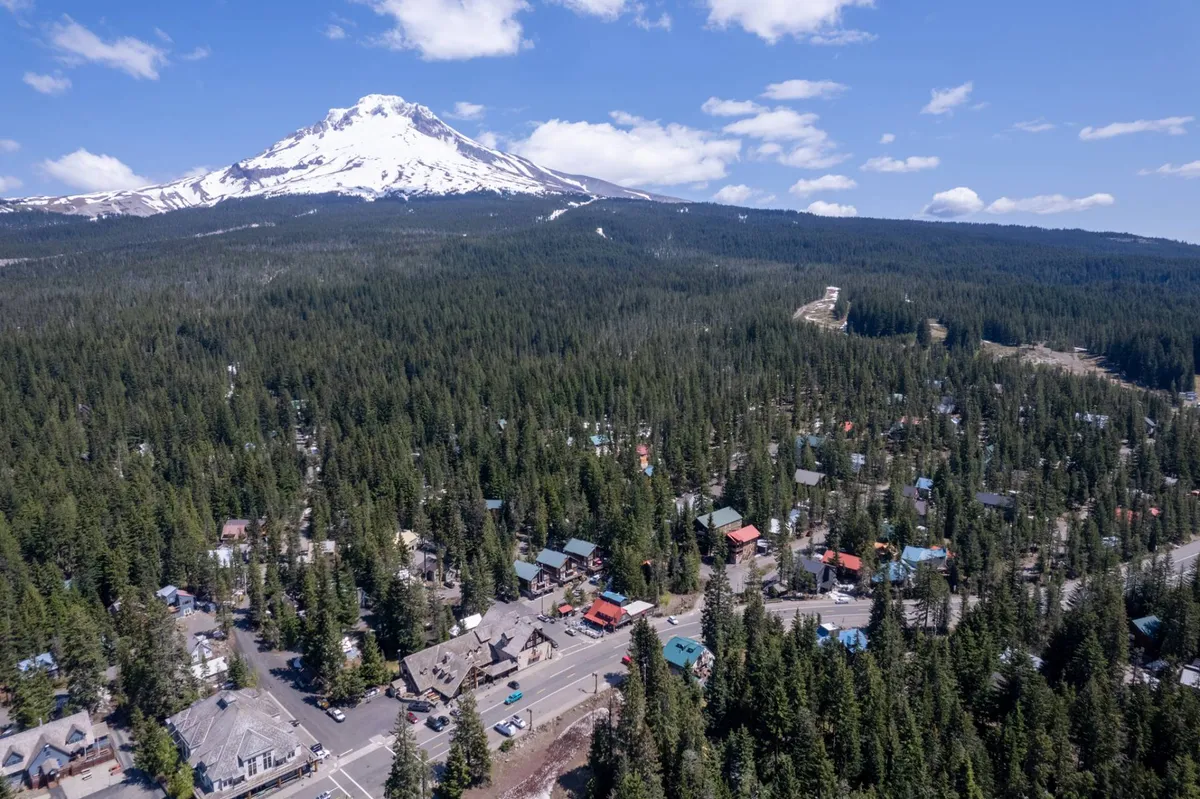 A breathtaking view featuring a snow-capped mountain rising above a dense green forest, with a small town nestled below and a scenic road winding through the landscape. The image feels both adventurous and peaceful, highlighting the proximity of natural wonders and community elements.