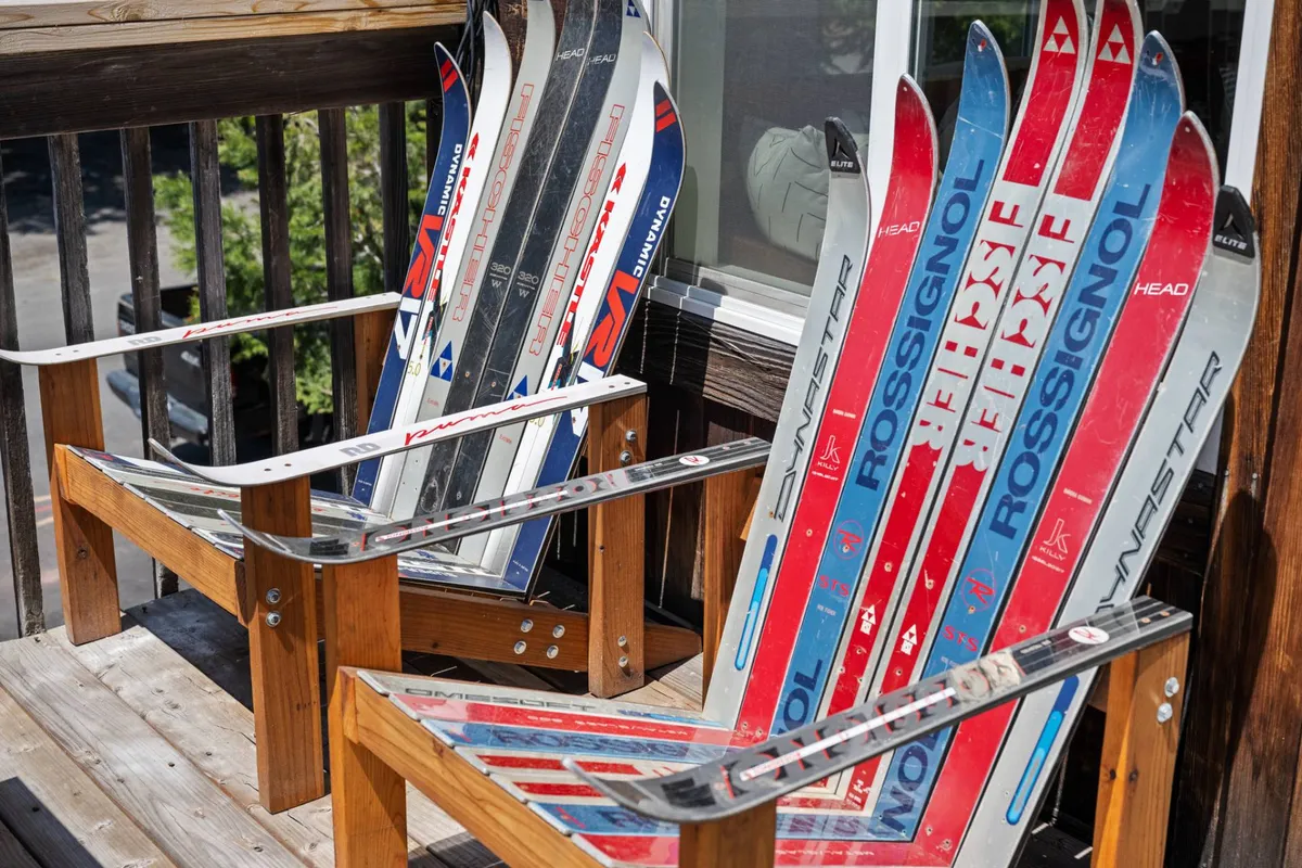 An exterior shot of a wooden deck framed by a sturdy railing, with playful custom ski chairs facing outward. Skis lean against the railing, while the surrounding pine trees provide a natural alpine backdrop. The atmosphere feels inviting and adventurous, ideal for relaxation or socializing.