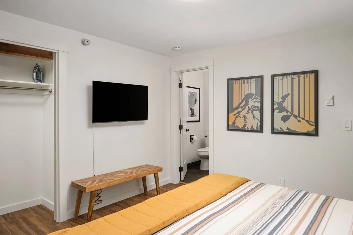 A cozy queen bedroom with soft yellow and white bedding, a wooden bench at the foot of the bed, framed artwork above the headboard, and a television mounted on the wall. The room is filled with natural light, creating a relaxing, homey atmosphere.