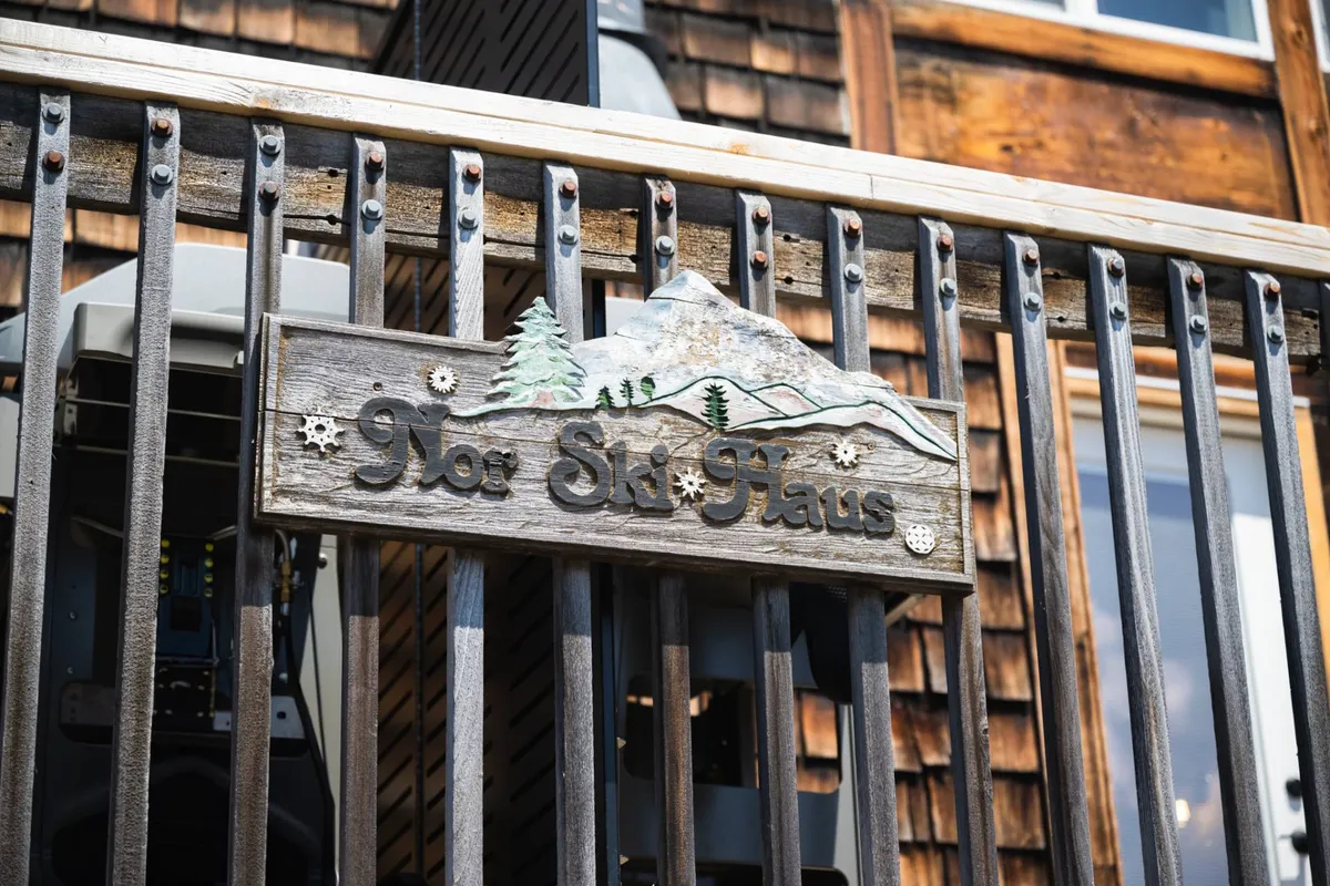 An exterior shot of a rustic wooden sign reading 'Not Ski Haus,' adorned with mountain and tree motifs and flanked by a cozy wooden railing. The image suggests a friendly, welcoming setting and hints at outdoor gatherings and the property’s mountain-lodge character.