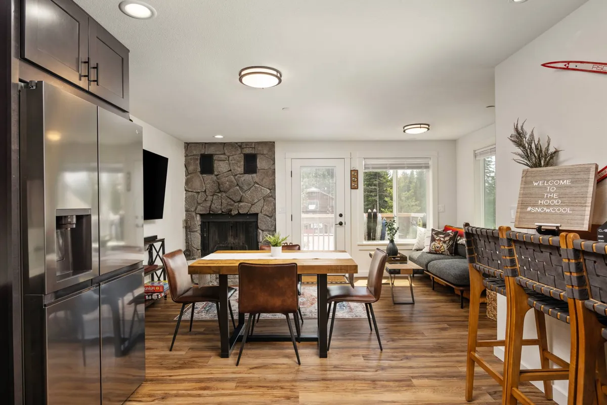 This image showcases a cozy, open-concept living and dining area featuring a prominent stone fireplace, a spacious dining table, bar seating, and large windows that allow natural light to flood the space. The inviting arrangement is perfect for gatherings, meals, and relaxing by the fire while enjoying views of the outdoors.