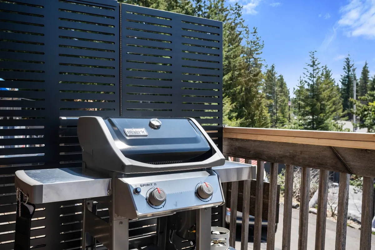 An outdoor deck scene highlighting a modern BBQ grill set against a wooden railing and decorative screen. Trees frame the background, creating a private, relaxed atmosphere for social gatherings or group feasts under the open sky.