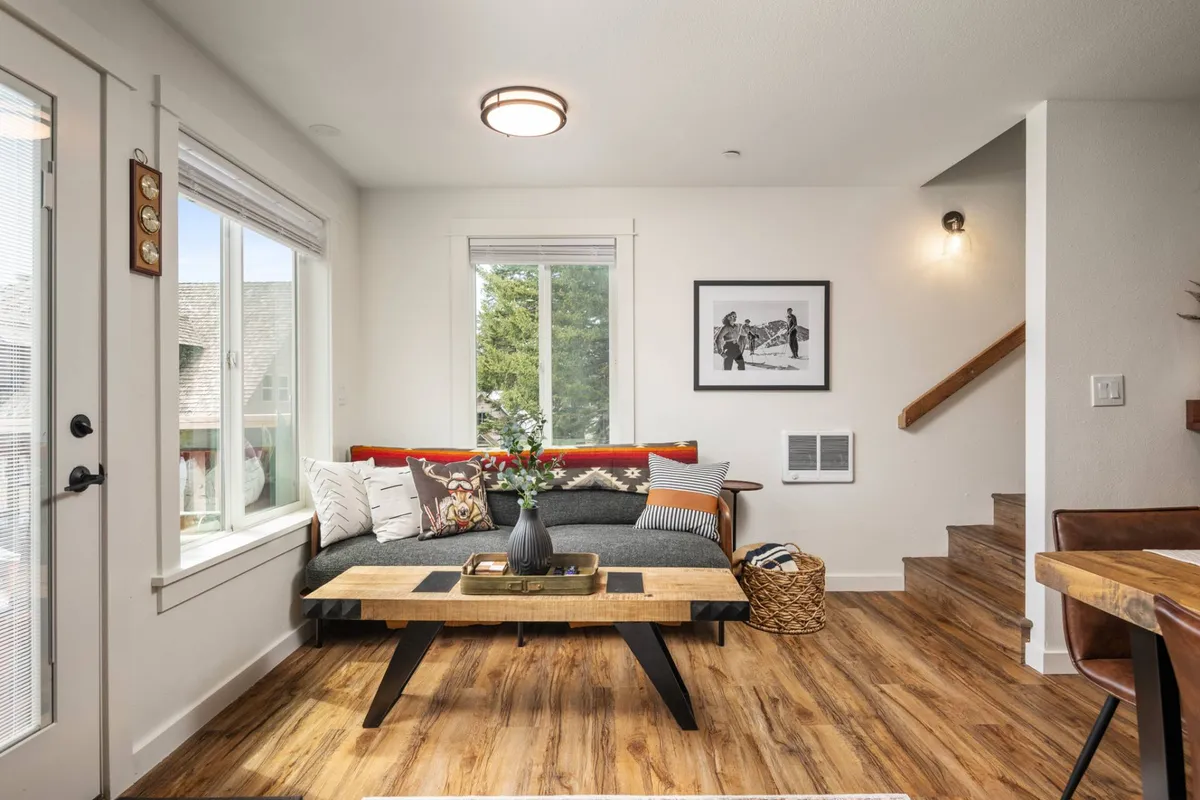 A welcoming living space with a plush sofa adorned with decorative pillows, a wooden coffee table ideal for gatherings, framed artwork on the wall, and ample natural light streaming in from a window. The staircase leading to the upper level hints at a spacious, multi-level layout.