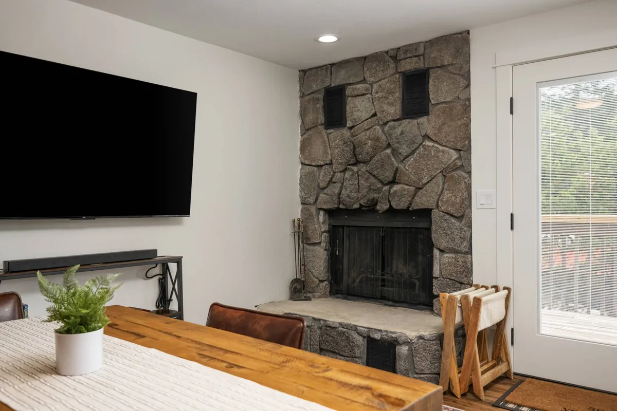 A welcoming living space with a large stone fireplace at its center, complemented by a big screen TV and a wooden dining table. The room has access to the outdoors, and the arrangement invites cozy evenings, movie nights, and shared meals while admiring the landscape outside.